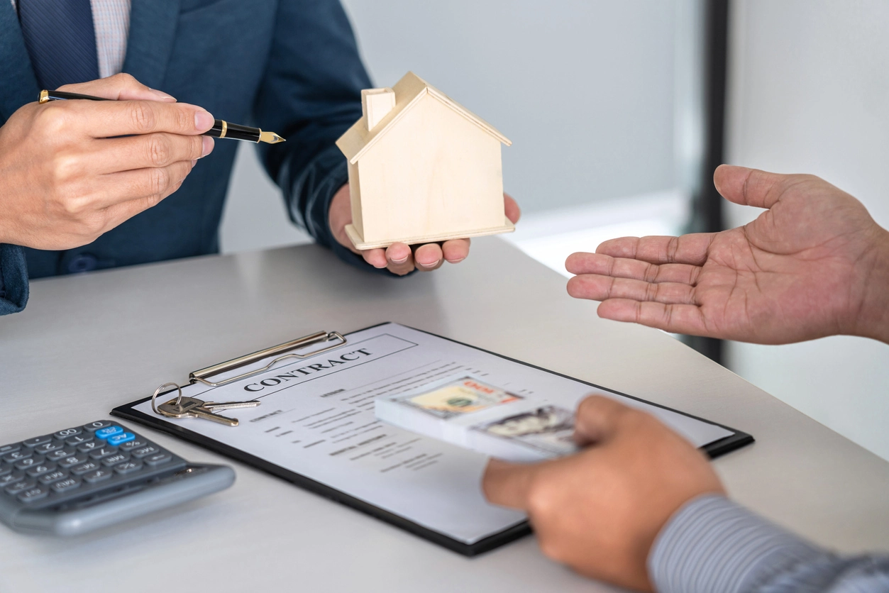 Deux personnes examinent un contrat de gestion locative, symbolisé par une maquette de maison, une calculatrice et des billets. L'image représente une situation liée à une faute du mandataire de gestion locative, comme une mauvaise gestion ou un désaccord contractuel