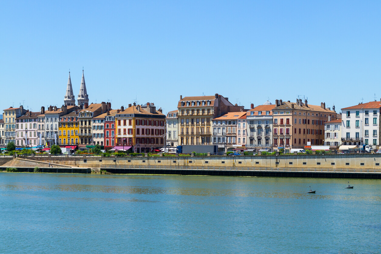 Une vue de bâtiments colorés le long de la Saône à Mâcon, en France. Les façades des immeubles sont dans des tons de jaune, rouge, beige, et rose, ajoutant de la couleur au paysage urbain. En arrière-plan, deux clochers se dressent, accentuant le charme architectural de la ville. Le ciel est dégagé, reflétant une journée ensoleillée.