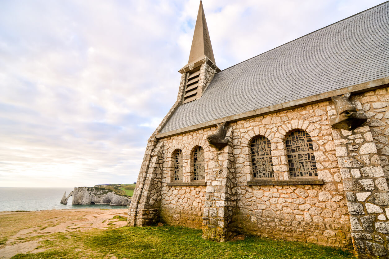 Image d'une chapelle en pierre située sur les falaises d'Étretat, en Normandie, avec des fenêtres en arc et un clocher pointu s'élevant vers le ciel. La scène se déploie avec en arrière-plan la mer et les falaises escarpées, capturant la beauté sereine du paysage côtier sous un ciel partiellement nuageux.