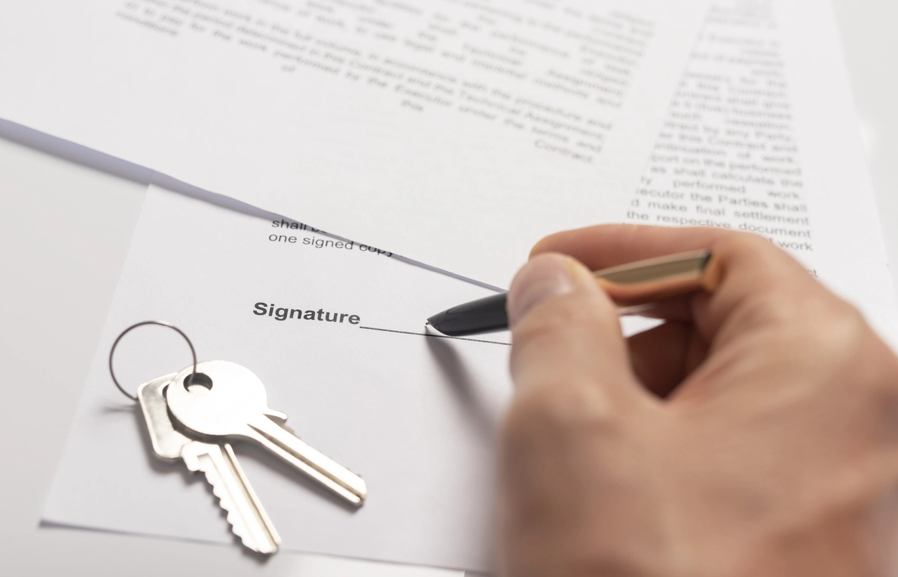 Une main signant un contrat de bail locatif avec un stylo noir, accompagnée d'un trousseau de clés posé sur le document, symbolisant la gestion d'un appartement locatif