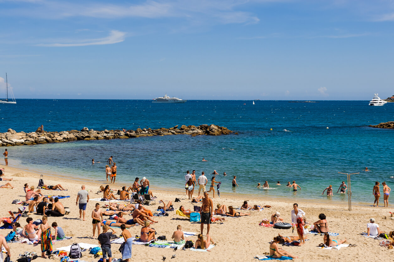 Une plage à Antibes ensoleillée avec de nombreuses personnes profitant du sable et de l'eau. Certains se prélassent sur des serviettes, d'autres discutent ou jouent dans l'eau. À l'arrière-plan, plusieurs yachts et voiliers flottent sur l'eau bleue de la mer sous un ciel clair avec quelques nuages blancs.