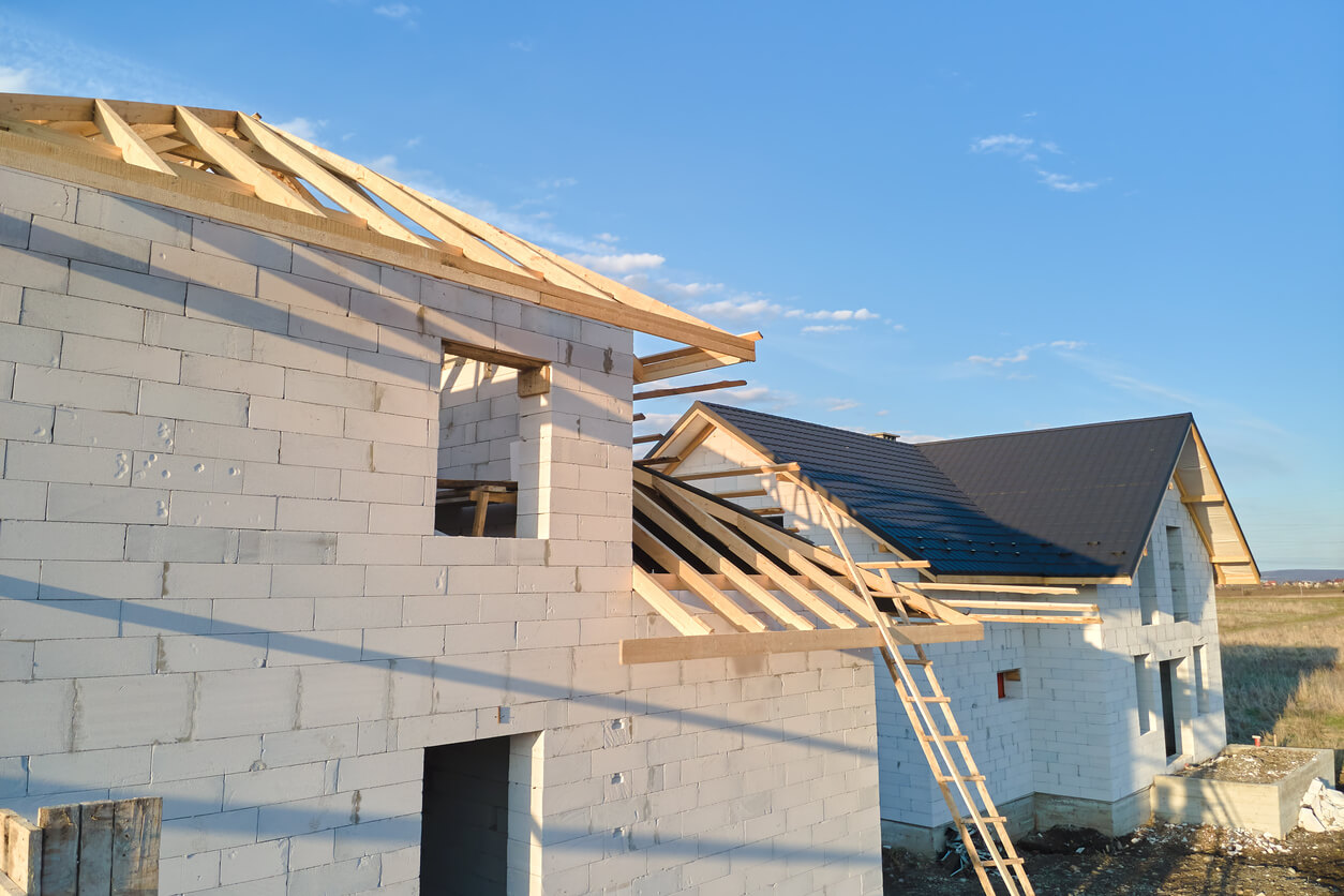 Une maison en construction, avec une échelle appuyée contre elle. Les murs sont en blocs de béton et le toit est en bois. Le ciel est bleu et il y a des nuages blancs.