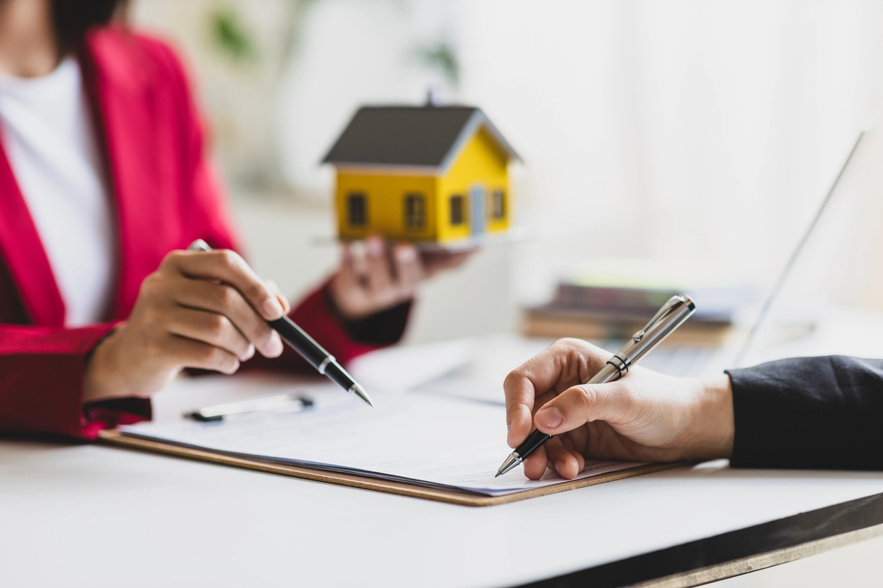 Une personne en costume signe un contrat de gestion locative posé sur une planchette. Une autre personne, vêtue d'une veste rouge, tient un stylo et une maquette de maison jaune