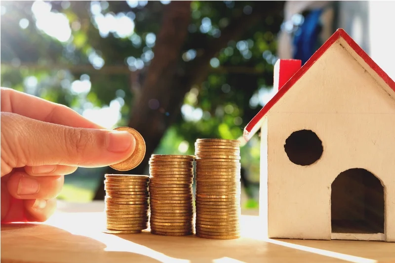 Une personne empilant des pièces de monnaie à côté d'une maquette de maison. Confier la gestion location maison à un professionnel optimise la rentabilité.