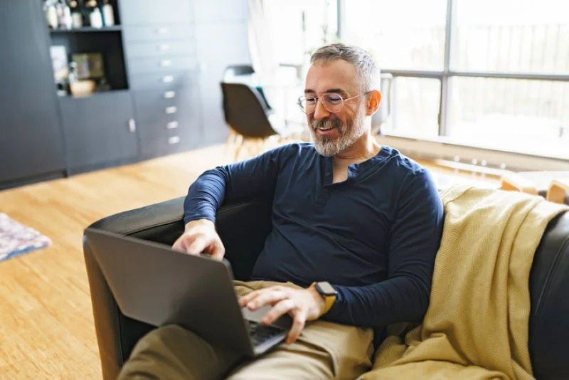 Un homme souriant entrain d'utiliser un ordinateur sur son canapé, dans une tranquillité d'esprit après un changement de gestion locative en cours de bail.