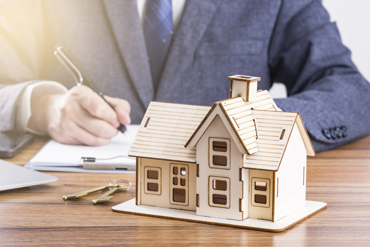 Un modèle réduit de maison en bois posé sur une table, accompagné de clés et d'un ordinateur portable, avec une personne en costume signant un contrat de la gestion locative Rennes en arrière-plan