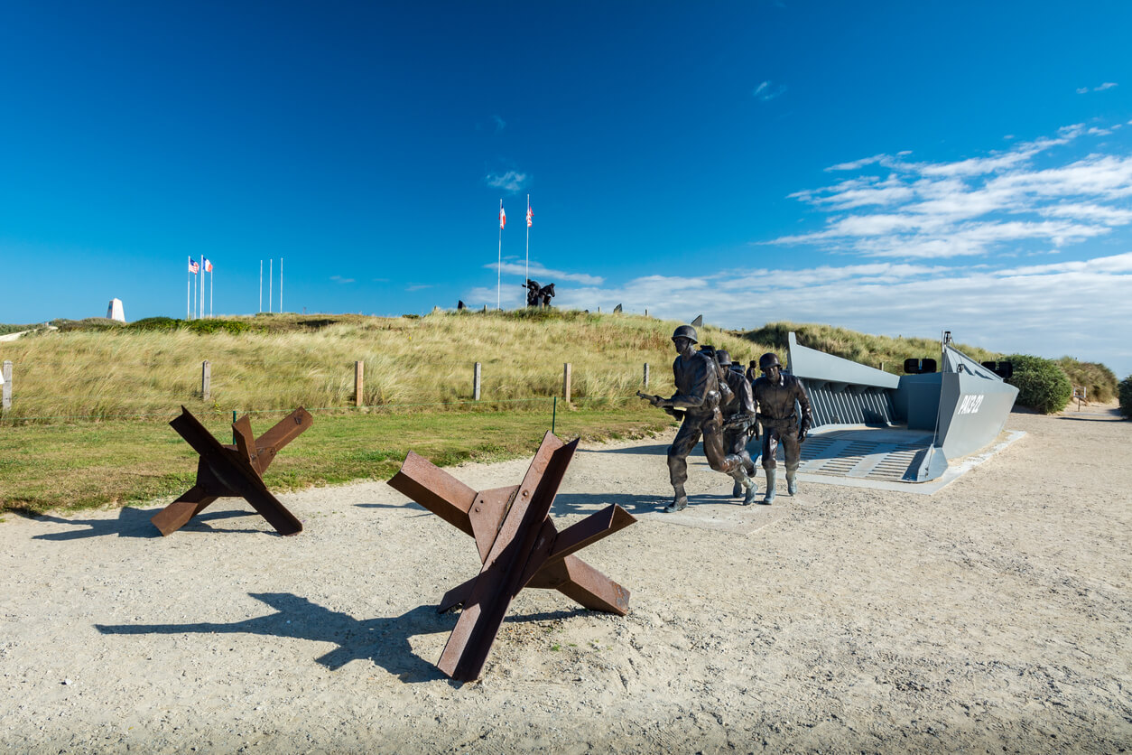 Un mémorial de la plage d'Utah Beach, en Normandie, France, représentant des soldats alliés en train de débarquer d'une barge de débarquement. Des statues de soldats et des obstacles antichars, tels que des hérissons tchèques, rappellent les défis auxquels les troupes alliées ont été confrontées. En arrière-plan, des drapeaux flottent au sommet d'une colline couverte de dunes, sous un ciel bleu dégagé.